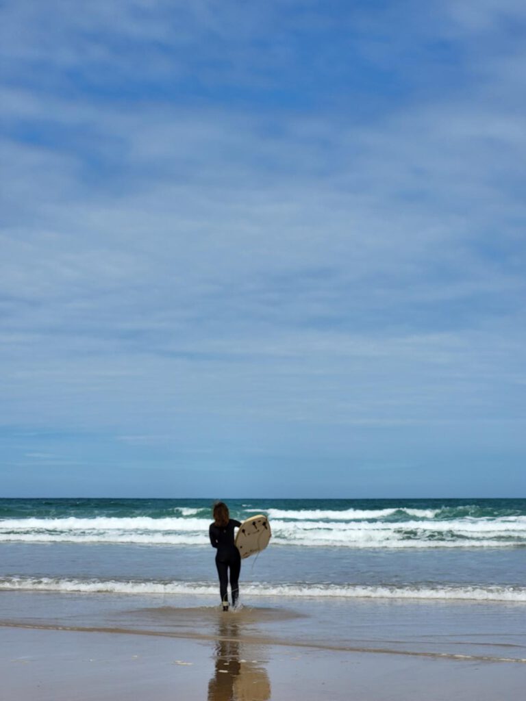 Eine Frau trägt ein Surfboard Richtung Meer.